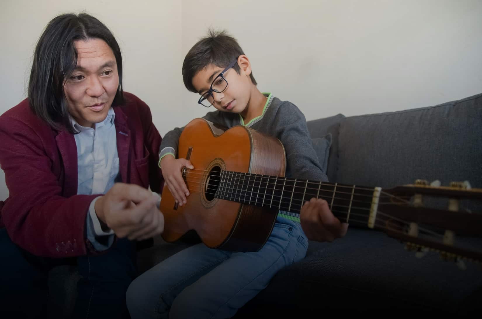 Boy with glasses receiving an acoustic guitar lessons from instructor on his left