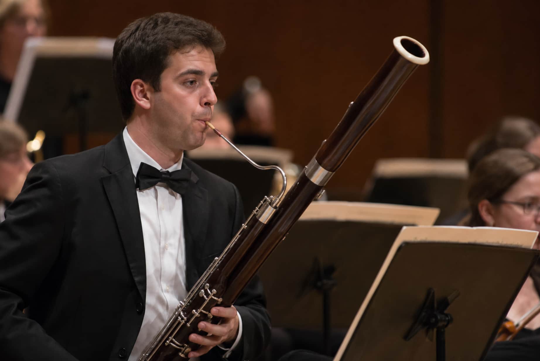 Close-up of a musician playing bassoon in a concert hall with an orchestra in the background.
