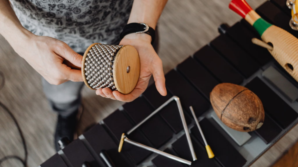 A teacher holding a cabasa surrounded by percussion instruments including a triangle, coconut shells and xylophone during percussion lessons
